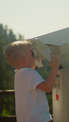 little boy with blond hair peers at mountains through enormous binoculars. child enjoys stunning view of sky and dense forest standing on top of mountain