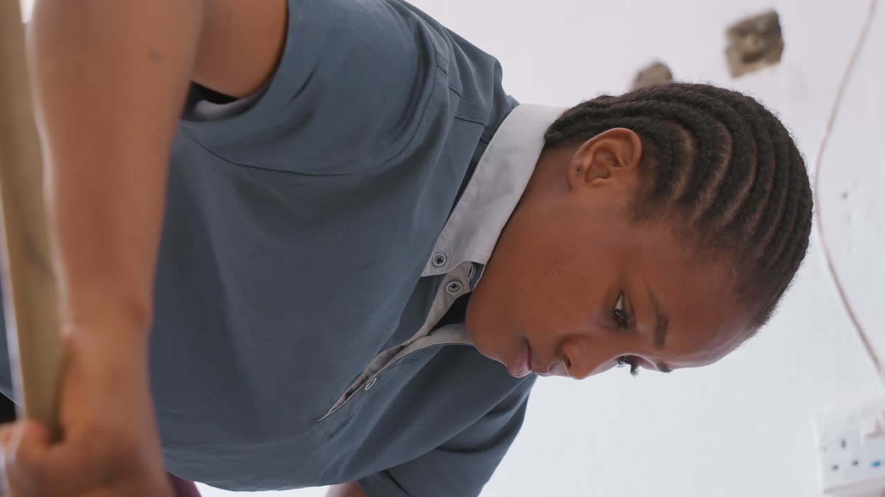 Black teen mopping house corridor intently, closeup profile of black girl in blue collared top, wet mop strokes near baseboard and outlet, concentrated cleaning motion, training as household helper