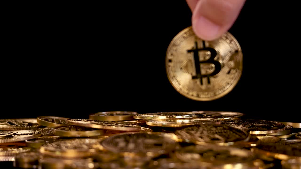 Hand flips golden Bitcoin token onto stack of coins, dramatic lighting, black background, close-up