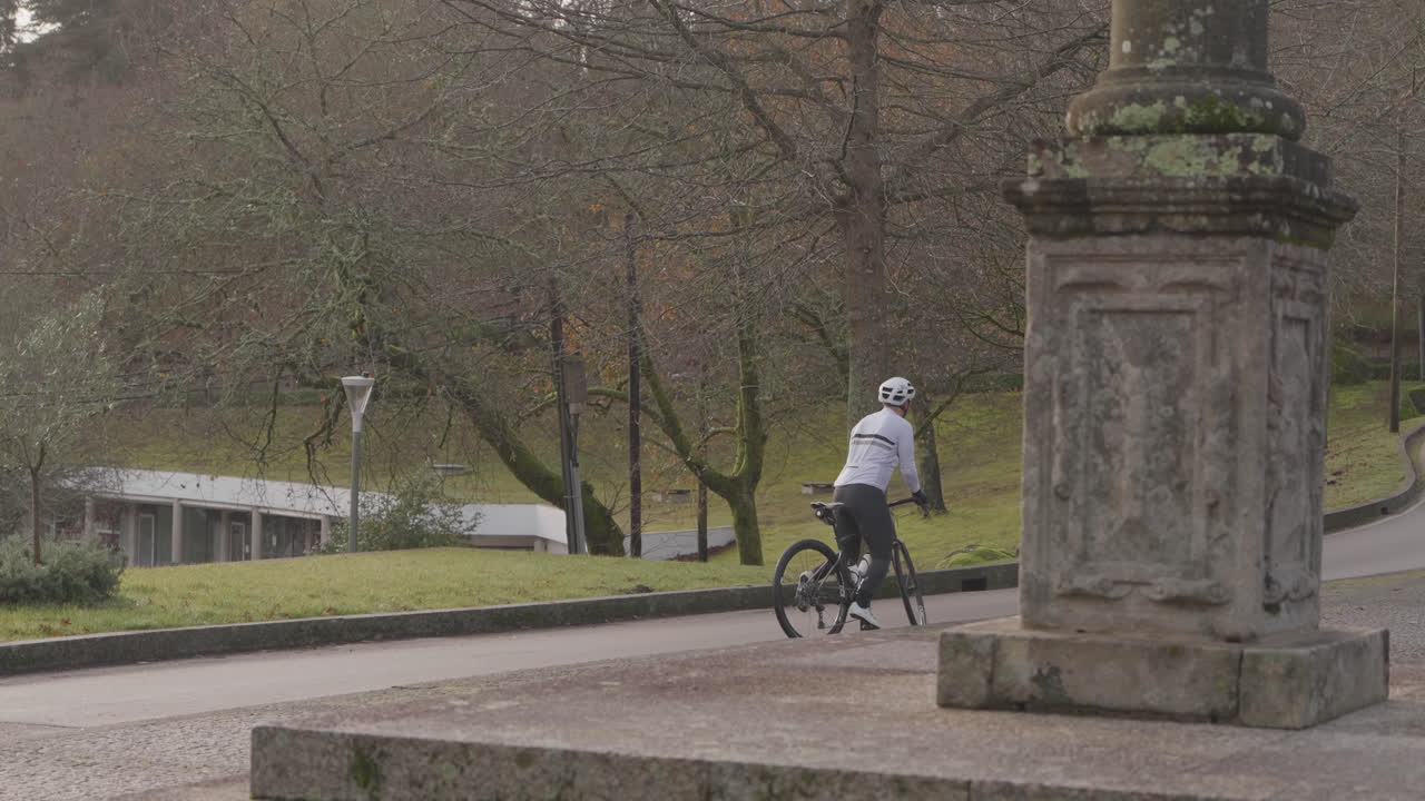 Persona in bicicletta su un sentiero asfaltato in un parco con alberi e una struttura in pietra