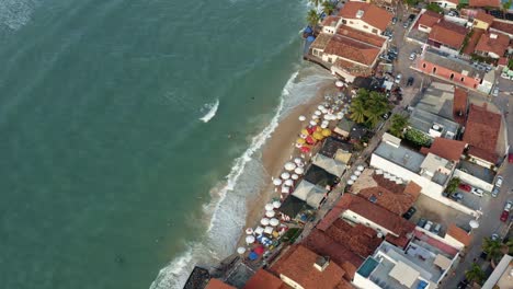 Rotierende-Luftdrohnenaufnahme-Aus-Der-Vogelperspektive-Des-Wunderschönen-Tropischen,-Berühmten-Pipa-Strands-Bei-Flut-Mit-Touristen,-Die-An-Einem-Sommerabend-Im-Wasser-Spielen-Und-Den-Schatten-Unter-Bunten-Sonnenschirmen-Genießen