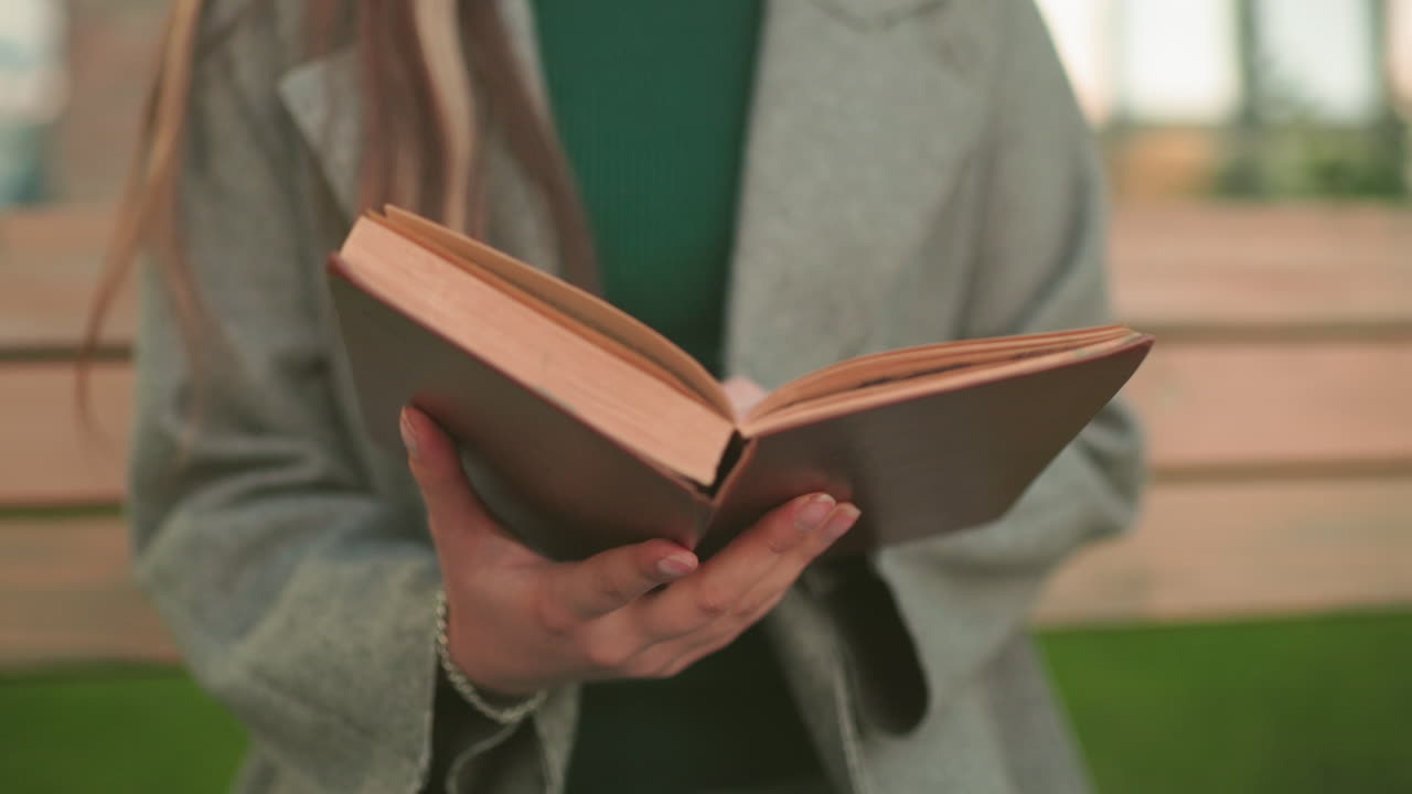 Close up of woman opening book outdoors while standing near wooden bench, hands holding pages with detail view highlighting reading, lifestyle, and study moment in natural setting