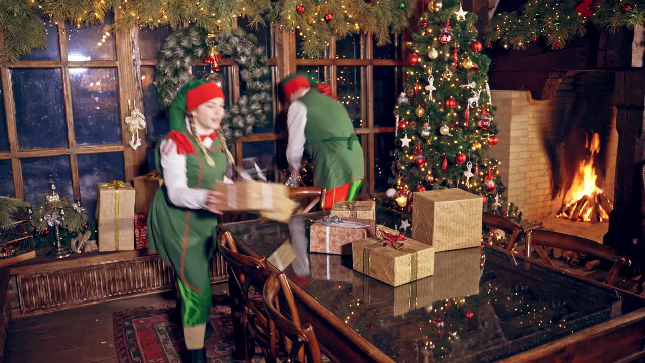 Two elves are standing in specially decorated room near the table, holding presents in hands. Feast fireplace in the background. Christmas spirit concept close up video. Christmas eve. New Year is coming. Elves are putting presents on the table.