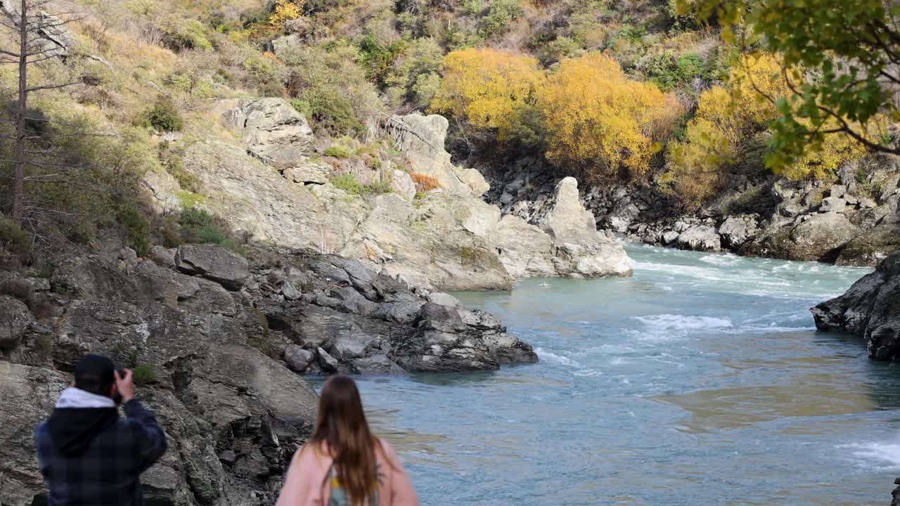 Two tourists observe the Kawarau River's flow amidst rocky terrain and autumn foliage in Queenstown, New Zealand