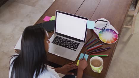 mujer de raza mixta usando una computadora portátil escribiendo en un cuaderno bebiendo café trabajando desde casa