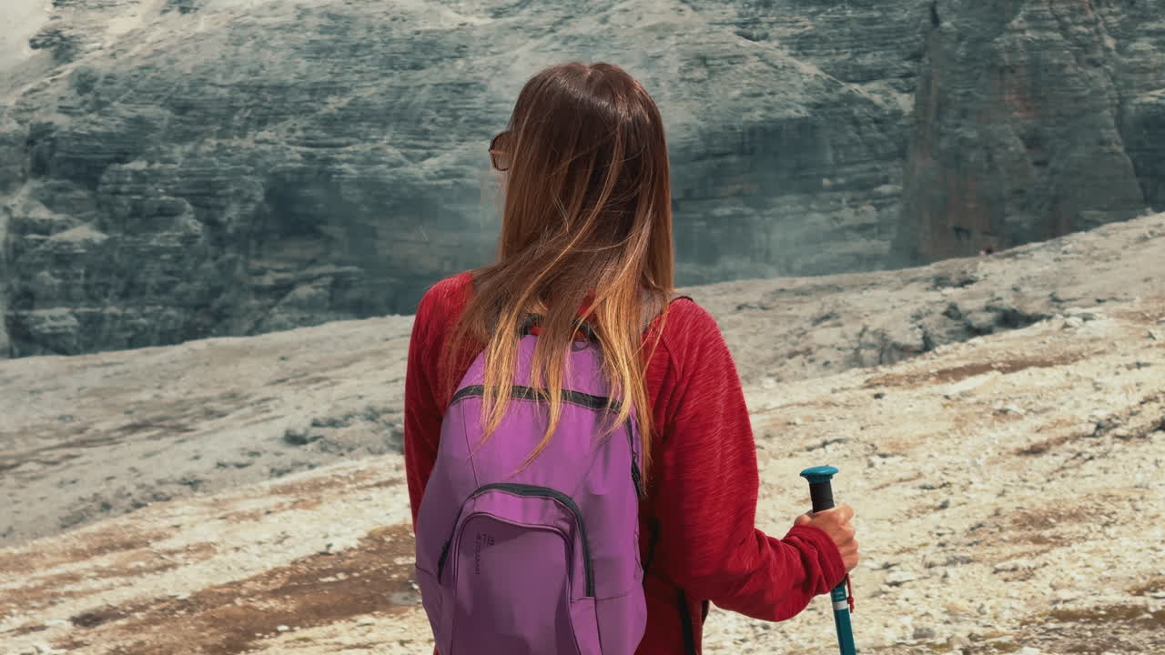 Female hiker standing on Pordoi Pass, directing attention toward rugged Sass Pordoi massif, surrounded by dramatic Italian Dolomites landscape during sunny summer day