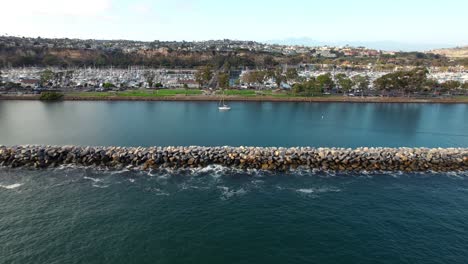 tracking shot along dana point jetty with sailboats on calm waters and cityscape background, california