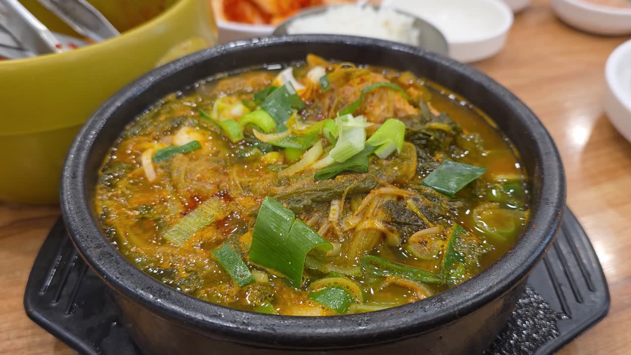 Close-up sup tulang punggung babi Gamja-tang yang mengepul dalam panci batu hitam dengan daun bawang, daun perilla, dan lauk kimchi di atas meja