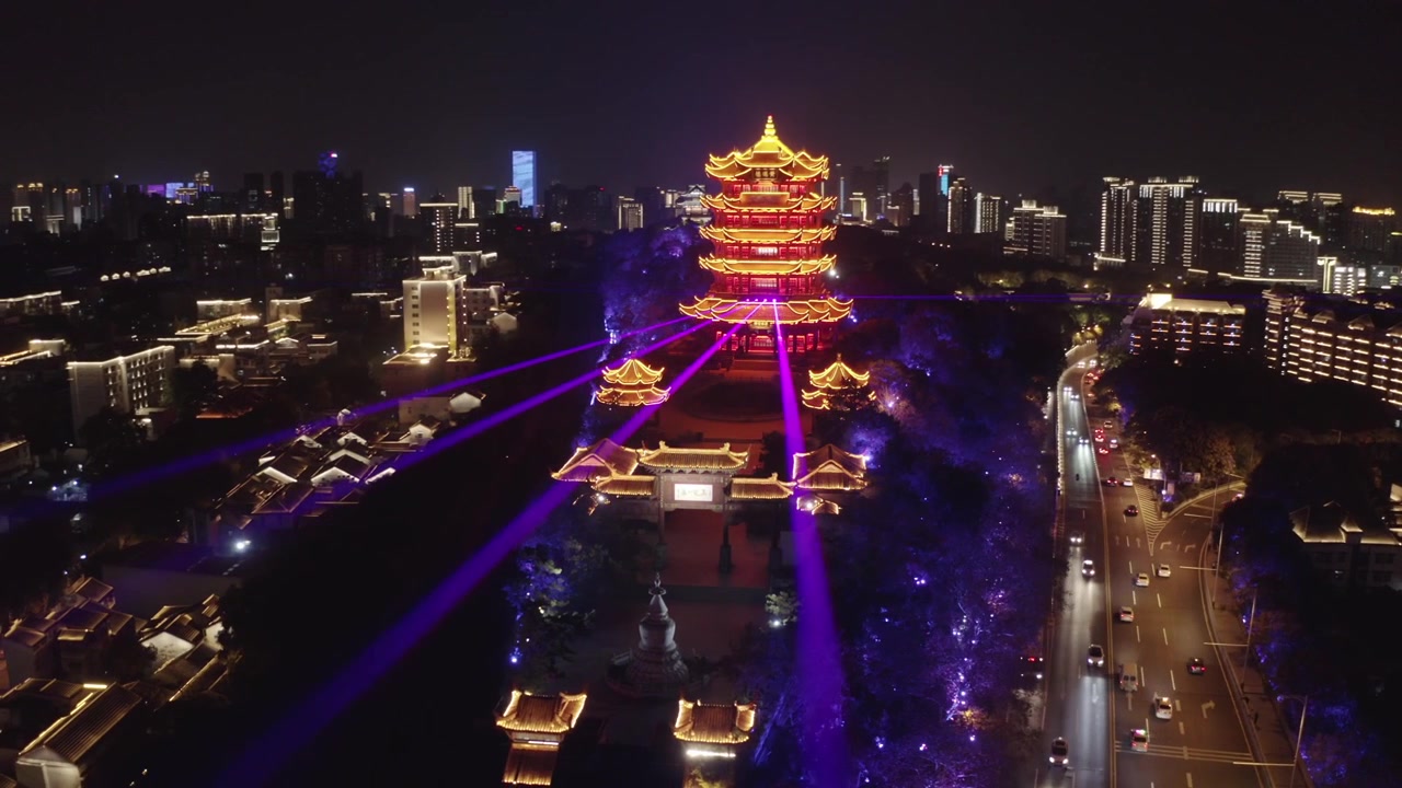Høy droneutsikt over det sterkt opplyste ikoniske Gule Kran-tårnet som gløder gyllenrødt om natten, omgitt av moderne bybygninger og trafikk i Wuhan, Hubei-provinsen, Kina