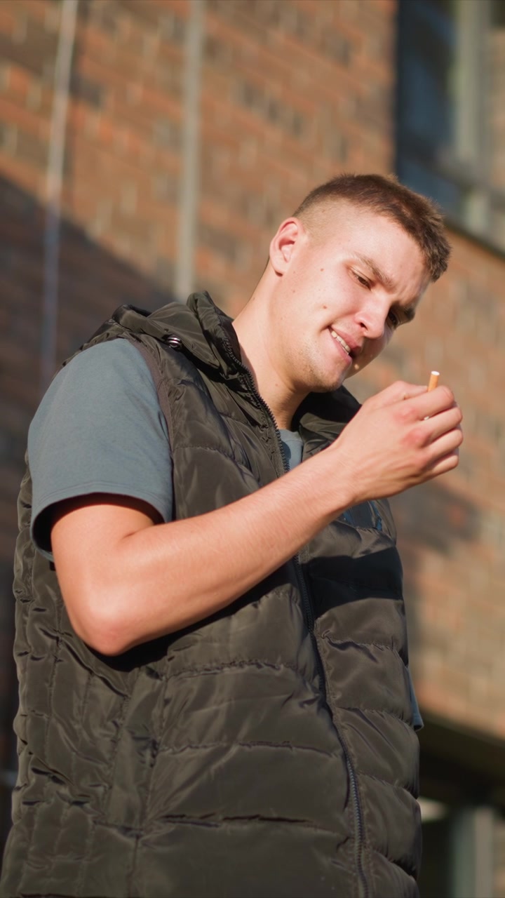 Close up of young man wearing dark puffer vest and gray t shirt standing outdoors in front of brick building smoking cigarette. Hand movements show him bringing lit end to his mouth then