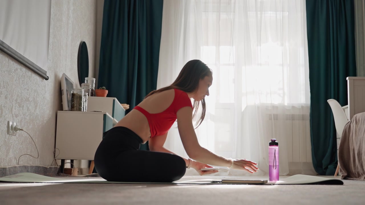 Donna che fa fitness a casa, si allunga sul tappetino, luce soffusa del mattino, interno della camera da letto, top sportivo rosso, leggings neri, capelli intrecciati, borraccia viola, che sistema il tappetino, che cerca idratazione, riscaldamento concentrato