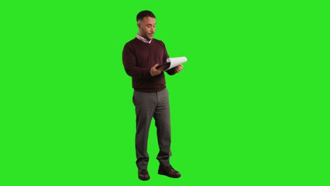 full length studio portrait of mature male teacher or businessman standing against green screen looking at clipboard