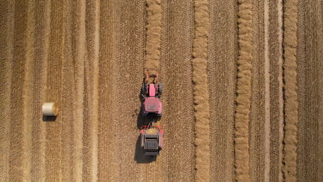 top down aerial - descending towards a tractor with a baling machine standing in a field next to a huge bale of straw