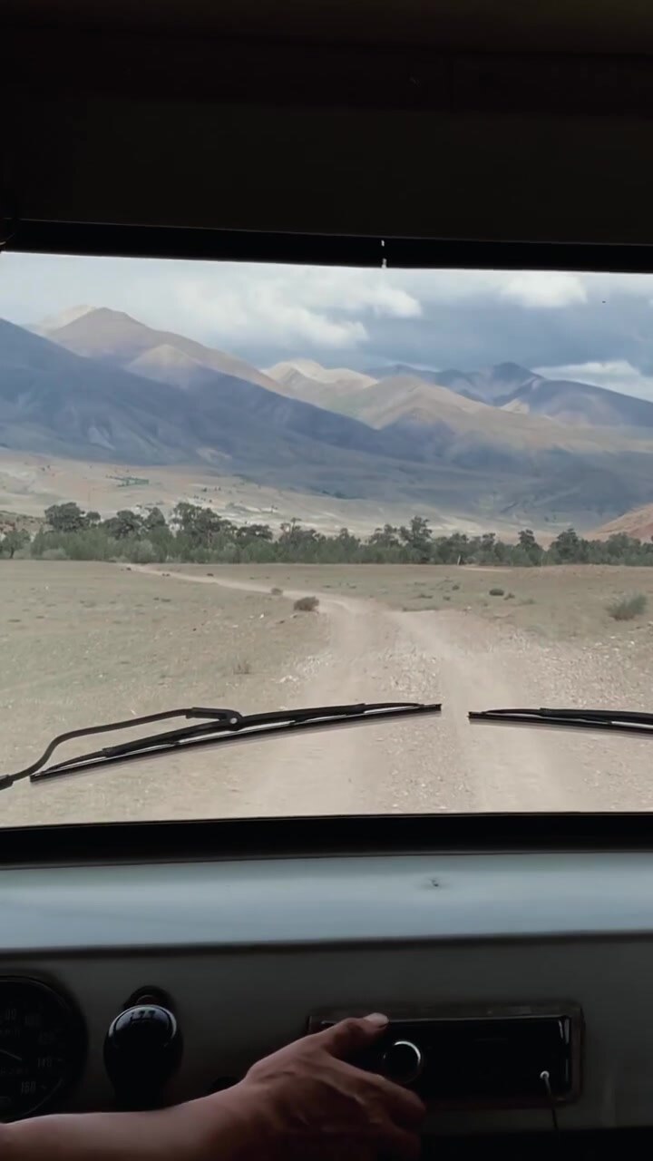 POV from inside a vehicle driving on a dusty dirt road across a wide valley with mountain range and clouds in the Altai Mountains, Altai Republic, Russia