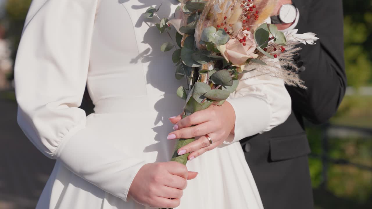 Detaljeret udsigt af en brud i elegant hvid kjole med en buket i hånden, med brudgommen i baggrunden. Den solbeskinnede have skaber en romantisk, fredfyldt atmosfære. Ideel til bryllupper