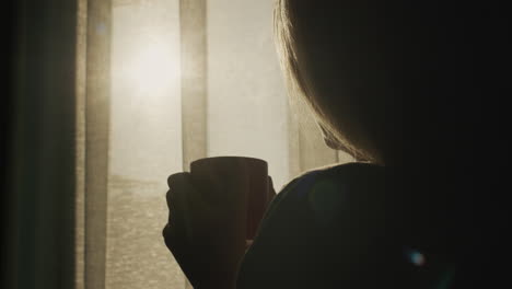 back view: a woman with a cup of hot tea looks out the window where the sun has risen. hot drink and good mood