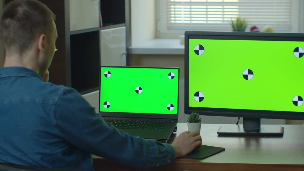 el editor de video masculino trabaja en su computadora personal con dos monitores de pantalla verde.