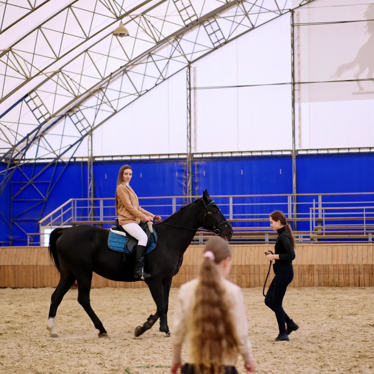 Long-haired woman riding a beautiful black horse. Female lady coach walks in front of animal. Young girl comes up to the foreground