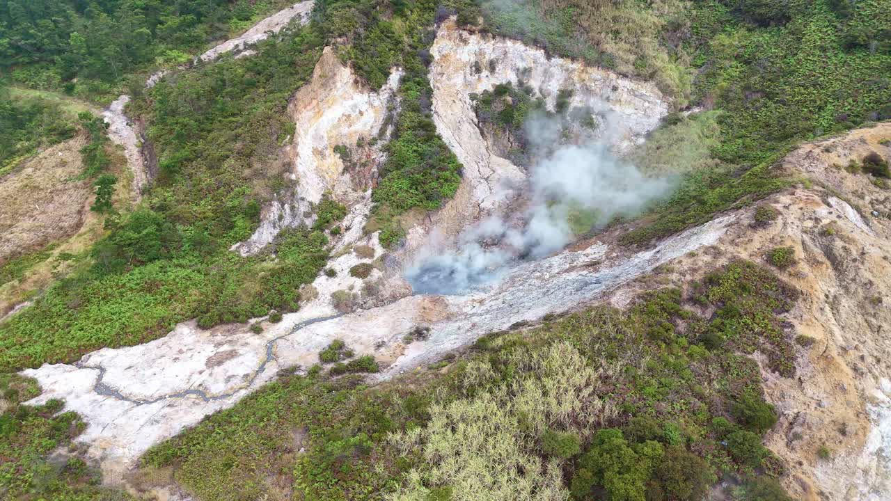 Aerial view of an active sulfur crater emitting steam and volcanic gas, surrounded by rocky terrain and green vegetation in a geothermal area