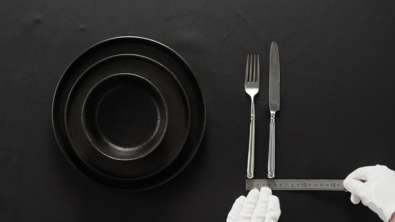 Minimalist table setting with white plate, fork, and knife arranged neatly, and a gloved hand adjusting cutlery.