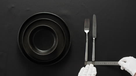 minimalist table setting with white plate, fork, and knife arranged neatly, and a gloved hand adjusting cutlery.