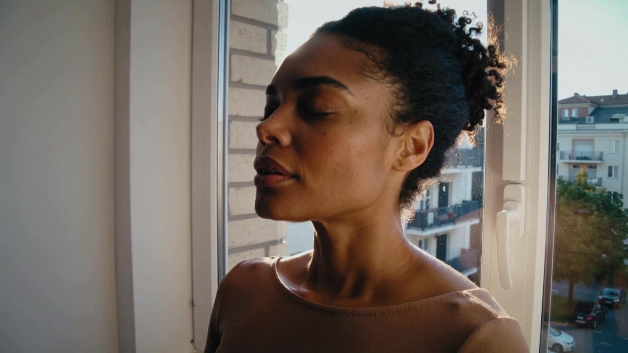 Sentendo la luce del sole, donna con chignon e top beige che chiude gli occhi, inclinando la testa alla finestra.