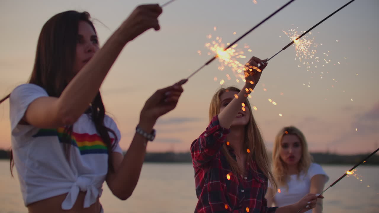 Three girls with nude waist are dancing with big bengal lights on the river coast. This is enjoyable summer evening on the open air party at pink sunset.