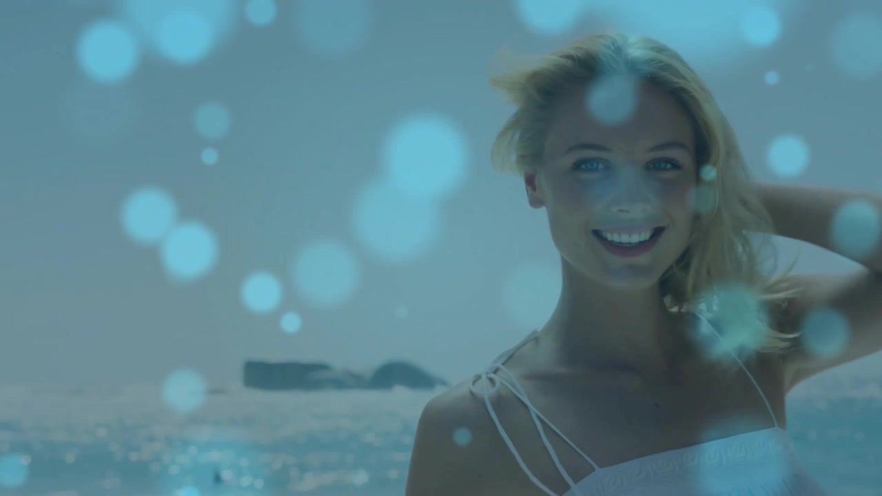 Modella in posa in spiaggia per un servizio di moda, si gira e sorride mentre un bokeh turchese fluttua.