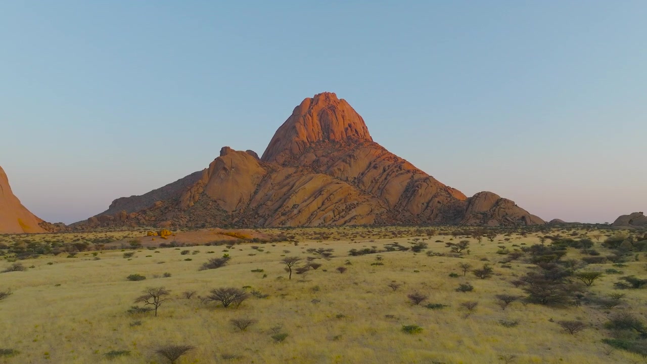A cinematic aerial tracking shot across the Namibian savannah towards the iconic Spitzkoppe granite peaks during a warm sunset. Ideal for travel documentaries and sustainability themes.
