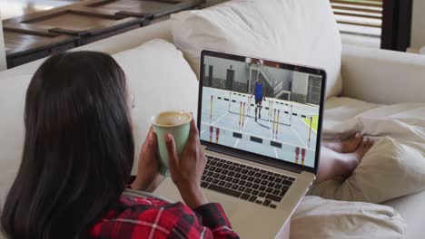 Composite-of-woman-sitting-at-home-holding-coffee-watching-athletics-running-event-on-laptop