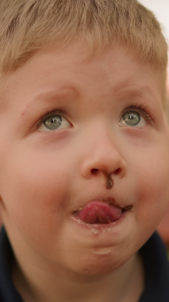 Funny toddler boy licks out chocolate from upper lip in city park at sunset closeup slow motion. Green eyed child with messy face. Sweet tooth in garden