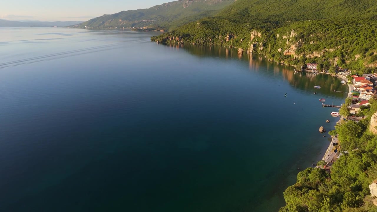 Ohri Gölü'nün devasa genişliğini sınırlayan kayalık ormanlık uçurumlar boyunca drone çekimi. Balkan vahşi doğa gölünün geniş yaz manzarası.