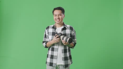 asian photographer holding a camera in his hands and smiling being happy while standing on green screen background in the studio