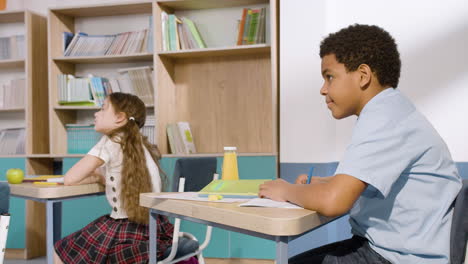 Little-Girl-And-American-Boy-Sitting-At-Desk-And-Taking-Notes-During-Class-At-School