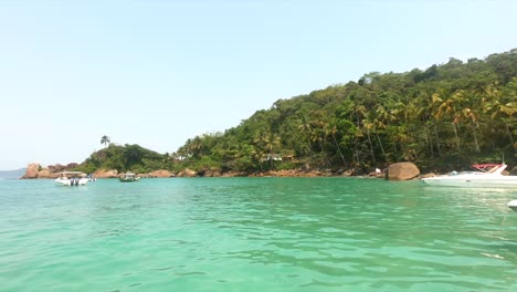 Tropical-island-with-blue-green-water-and-palm-trees,-boats-bobbing-in-front-of-blue-sky-and-rocks-in-Isla-Grande-Brazil