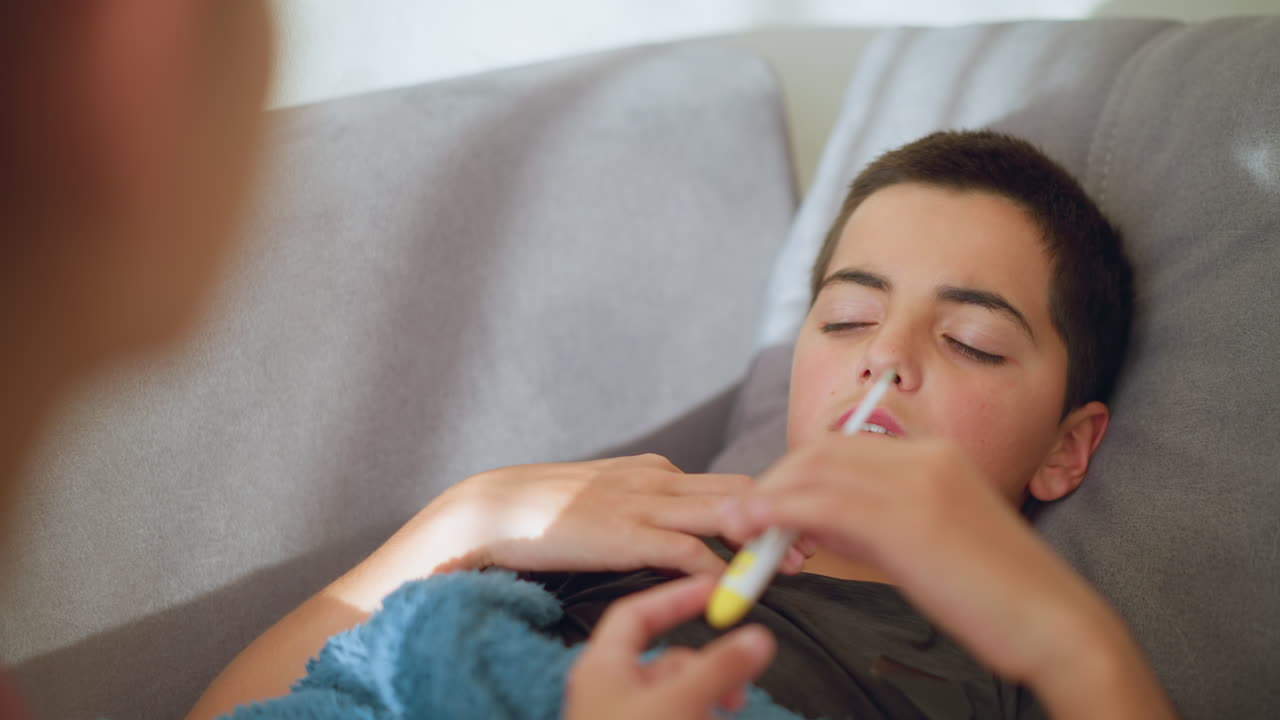 Primo piano di persona che porge un termometro a un bambino malato coperto da una coperta, bambino che riceve il termometro, espressione preoccupata, ambiente interno con illuminazione soffusa