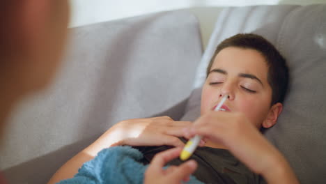 close-up of individual handing thermometer to sick boy covered with blanket, boy receiving thermometer, concerned expression, indoor setting with soft lighting