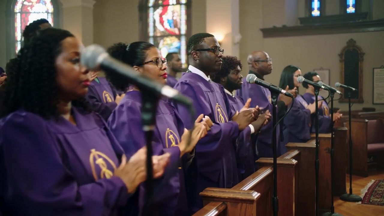 Cantanti del coro che applaudono e cantano lodi in chiesa, con la telecamera che si avvicina mostrando microfoni e vesti viola.