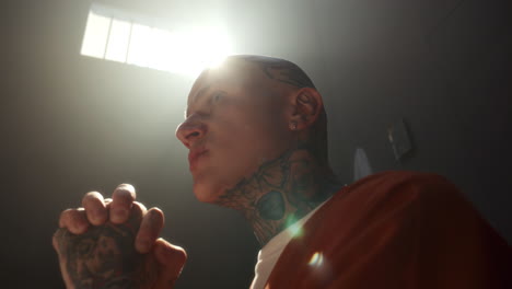 tattooed inmate saying prayer with hands clasped in sunlit prison cell