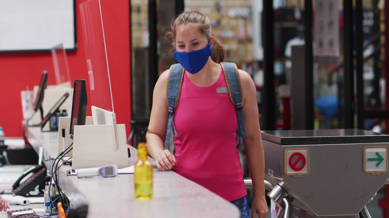 Mujer caucásica con mascarilla hablando en la recepción de un gimnasio y desinfectándose las manos