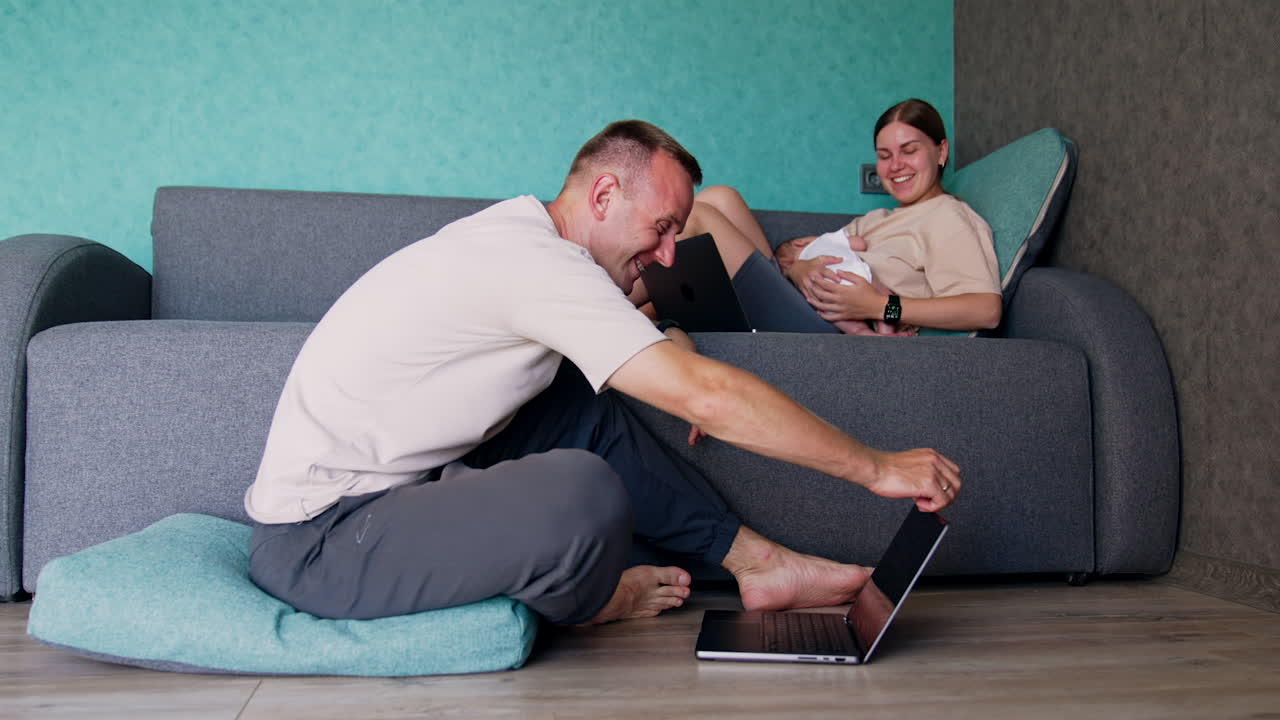 Man and woman working at home are laughing cheerfully. Parents work on laptops. Woman holding a baby in her hands.