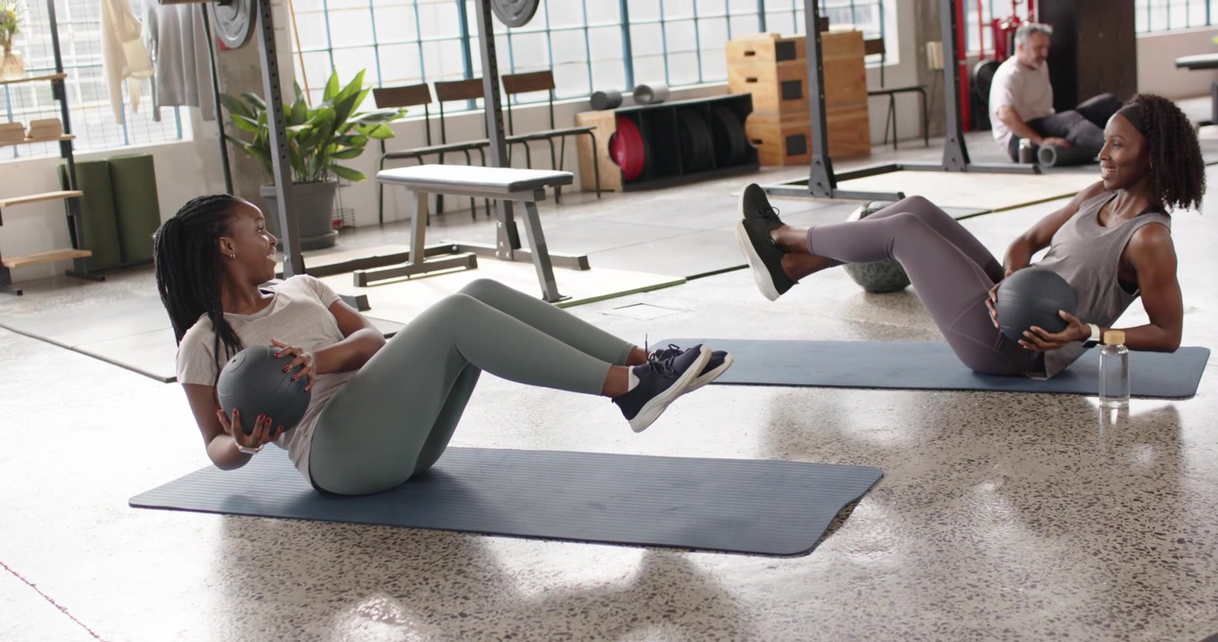 African American women in sportswear lifting feet twisting on gym mats with medicine balls for core