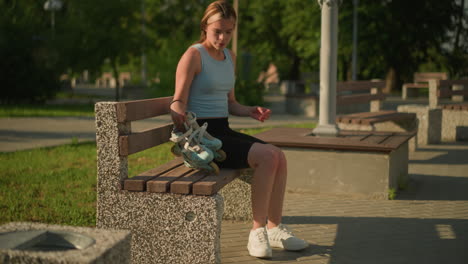 young lady placing cyan roller skates on wooden bench outdoors, she is wearing black shorts and white sneakers while background features trees, greenery, and multiple benches under bright sunlight