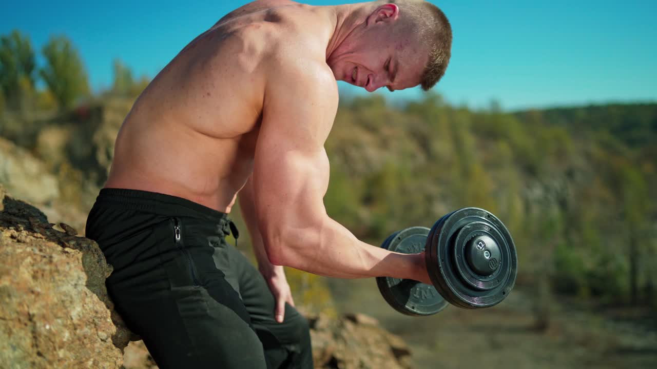 Sportsman pumping arm muscles on rocky background. Shirtless athletic man exercising with metal dumbbell in nature. Close-up.