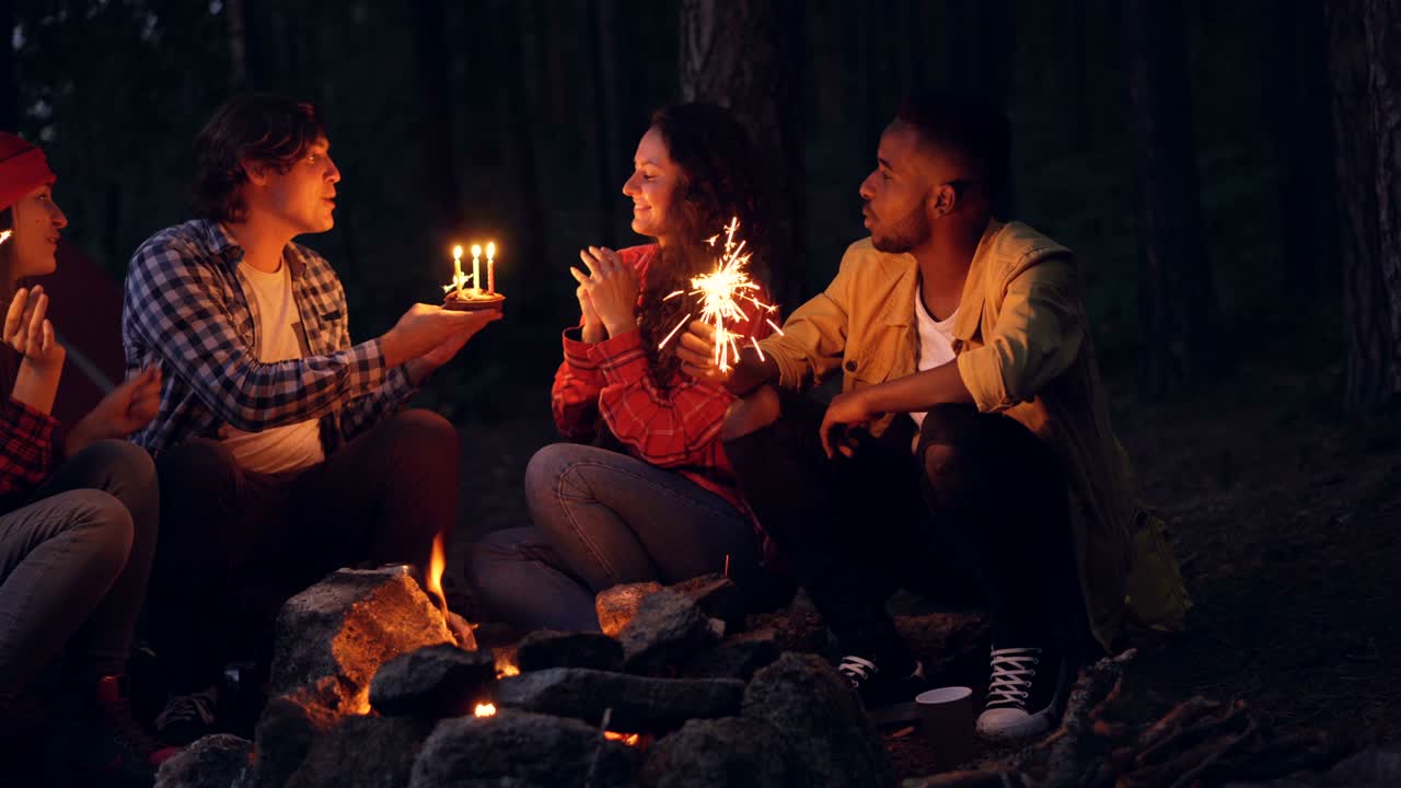Young woman is celebrating birthday in forest with friends blowing candles on cake and clapping hands while people around are holding bengal lights and congratulating her.