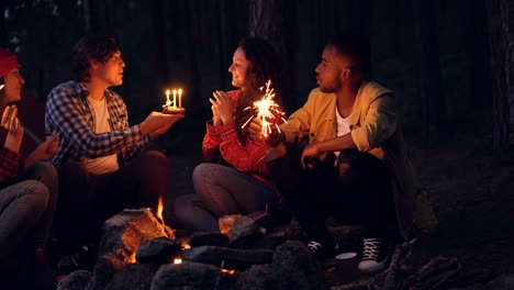 una joven está celebrando su cumpleaños en el bosque con amigos soplando velas en el pastel y aplaudiendo mientras la gente de alrededor sostiene luces de bengala y la felicita.