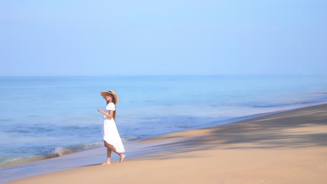 Eine-Junge-Frau,-Die-Ein-Fließendes-Weißes-Sommerkleid-Und-Einen-Strohhut-Trägt,-Geht-Am-Strand-Entlang-Und-In-Die-Ankommende-Brandung