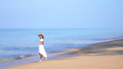 흐르는 흰색 선드레스와 밀짚 모자를 쓴 젊은 여성이 해변을 따라 다가오는 파도 속으로 걸어갑니다.