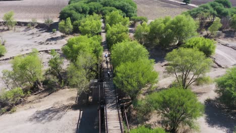 Group-of-friends-riding-bicycle-crossing-an-old-bridge-through-an-amazing-landscape,-drone-shot