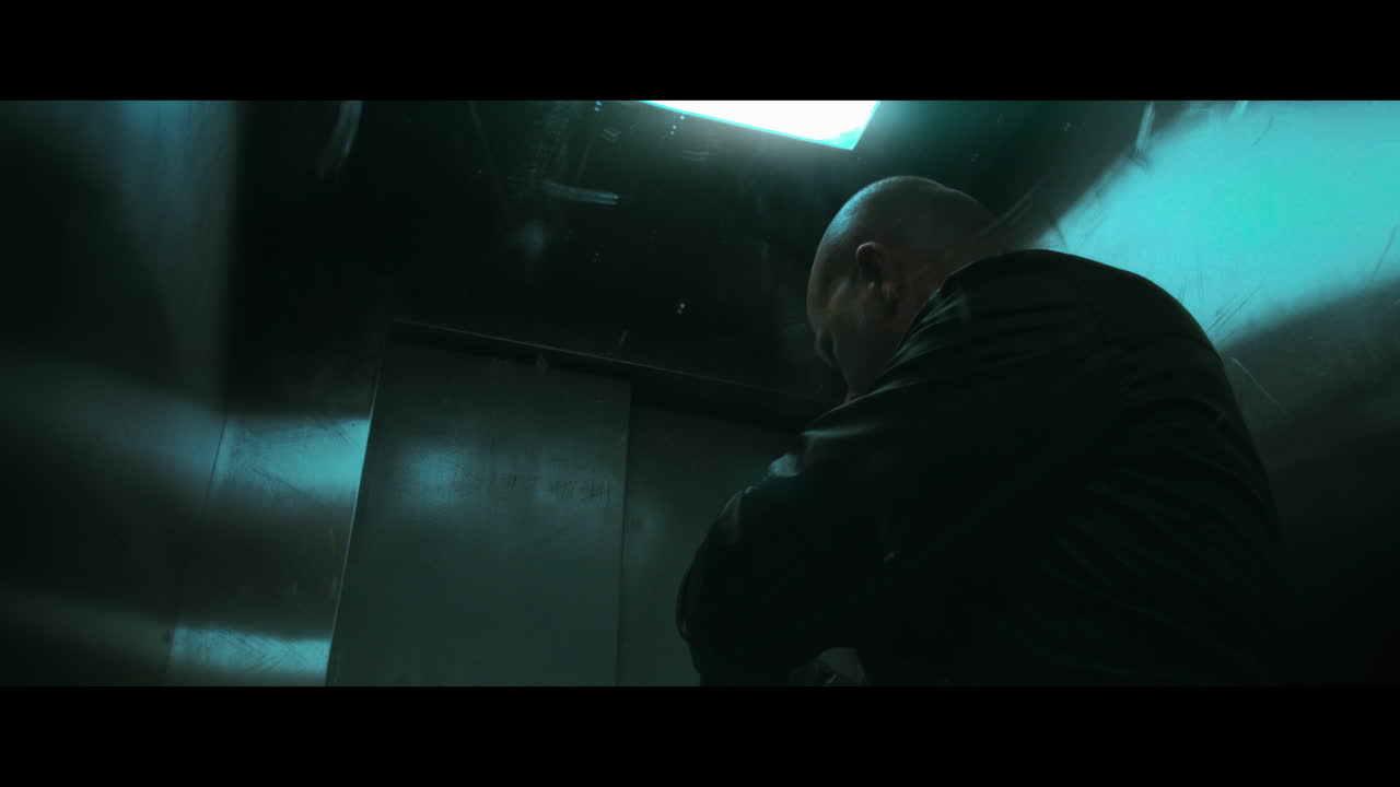 A bald dangerous looking man prepares a gun with the silencer in the elevator